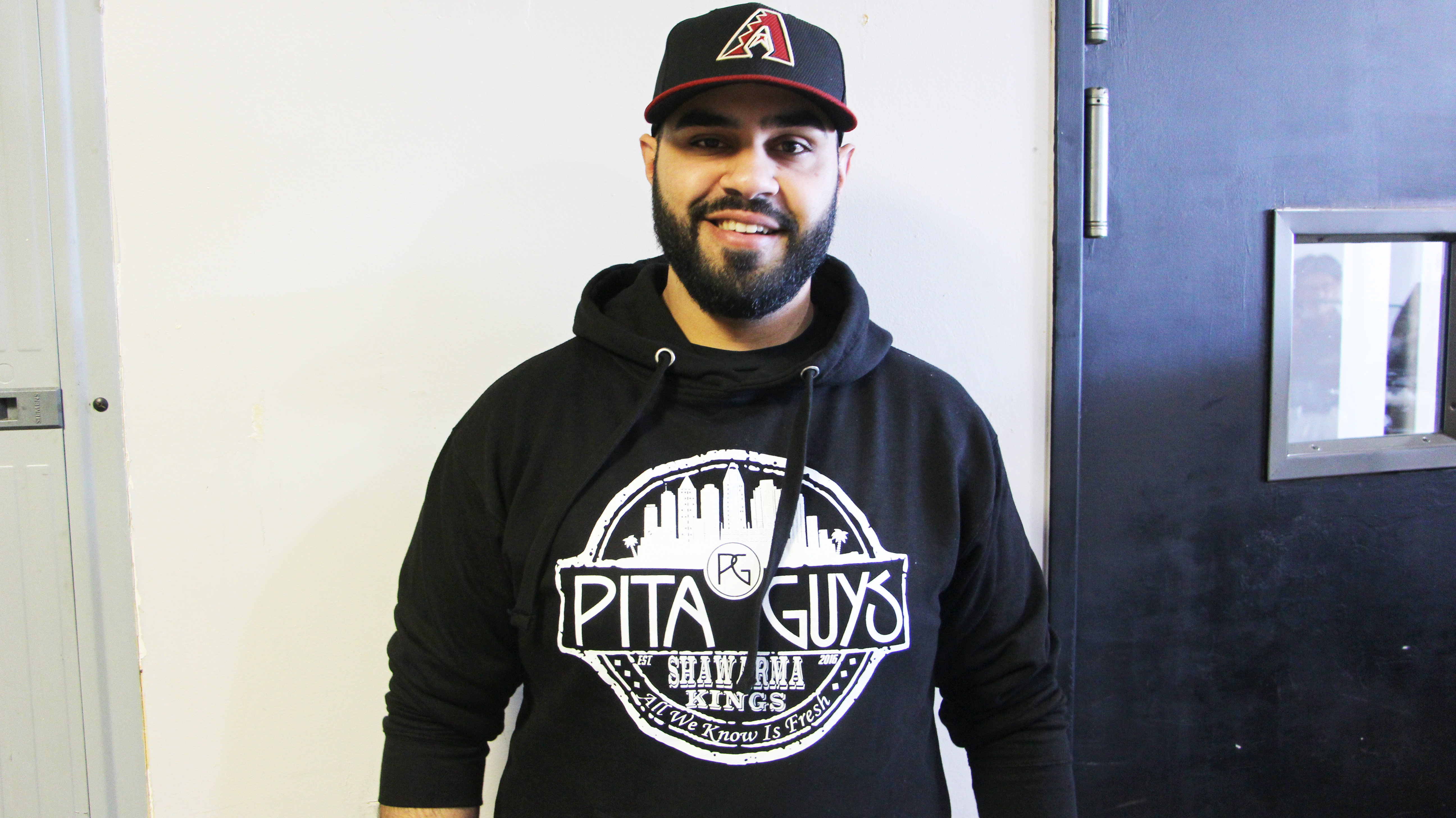 Man wearing a black hoodie with a Pita Guys logo and a black baseball cap.
